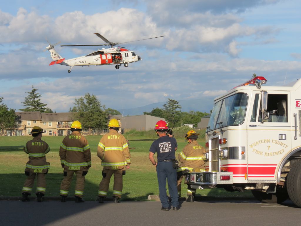 WCFD7 personnel watch as NAS Whidbey Island S&R crews simulate a rescue via helicopter at the Mountain View school campus (June 12, 2017). Photo: Whatcom News