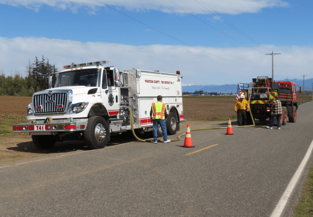 Crews from WCFD7 and WCFD14 work to recharge a brush truck at a 30-acre wildfire near the border on Bender Road (April 19, 2024). Photo: Whatcom News